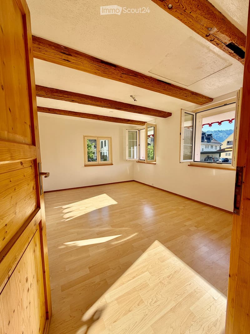 Empty room with wooden floors, exposed wooden beams, and multiple windows letting in natural light.