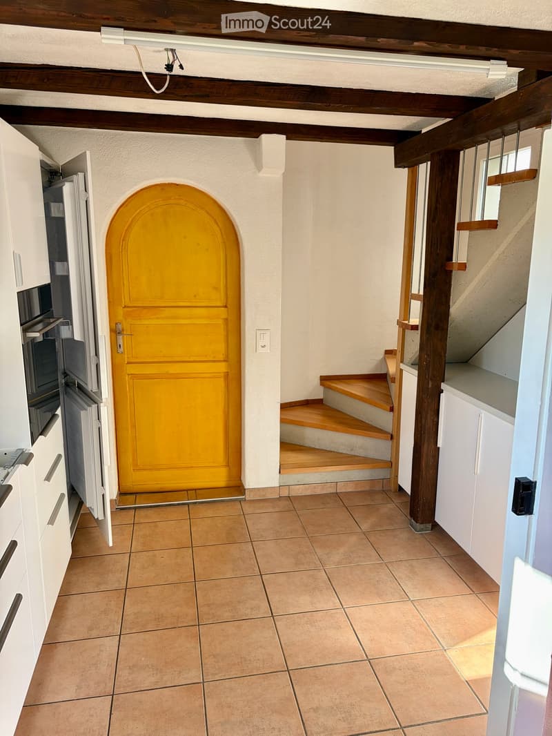 A narrow hallway with tiled flooring, white walls, and wooden beams on the ceiling. Features a bright yellow arched door, a small staircase with wooden steps, and built-in white cabinets on the left side.