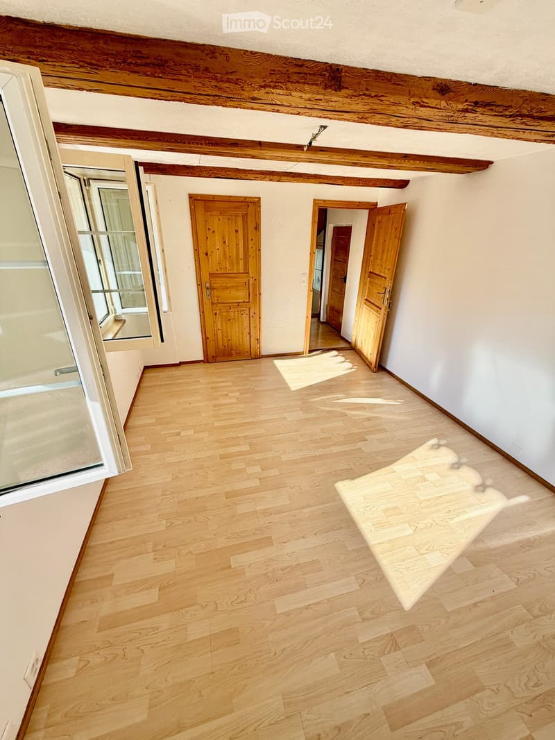 Empty hallway with wooden flooring, white walls, and exposed wooden beams on the ceiling. Features two wooden doors and a window with white frame.