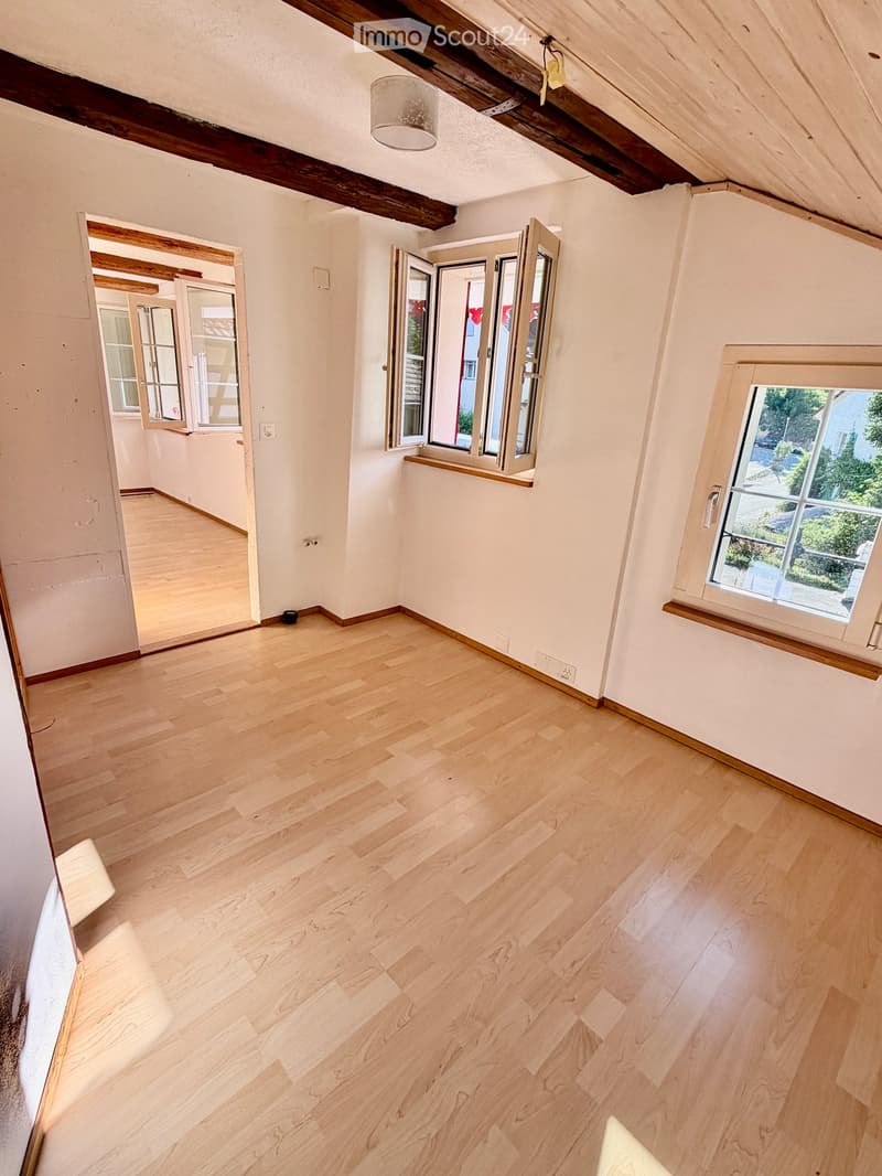 Empty room with light wooden flooring, white walls, and exposed wooden beams on the ceiling. Features multiple windows with open shutters and a visible doorway leading to another space.