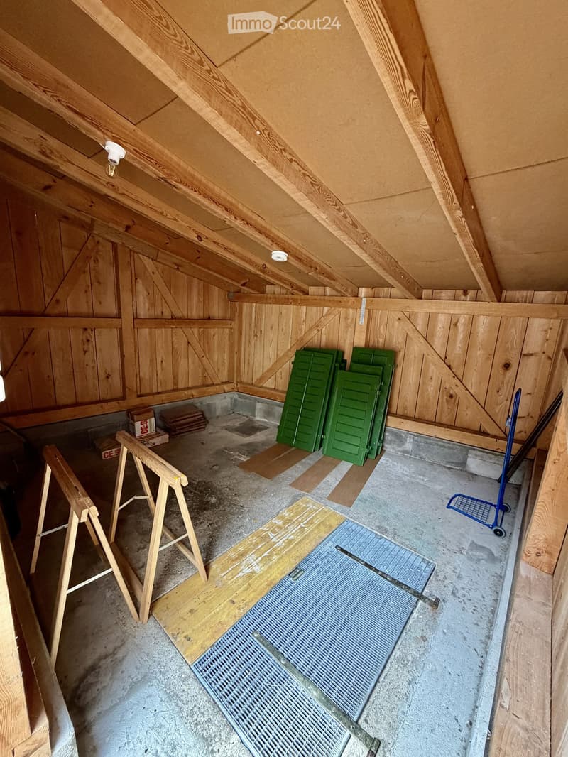 An unfinished attic space with exposed wooden beams, concrete floor, and various construction materials including wooden planks, green shutters, and a metal grate.
