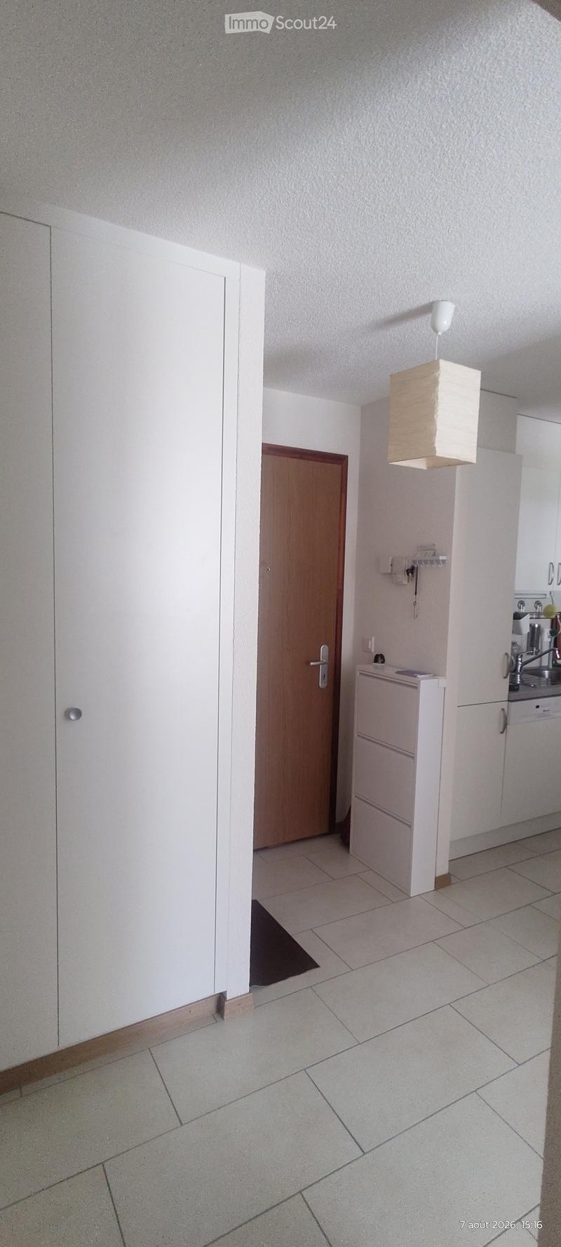 Empty hallway with tiled floor, white walls, a wooden door, and a small kitchen area visible in the background.