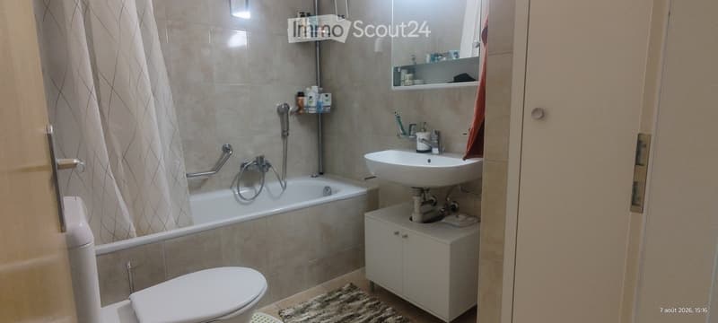 Bathroom with a bathtub, toilet, sink, mirror, and a small cabinet. The walls are tiled, and a towel hangs on the side.