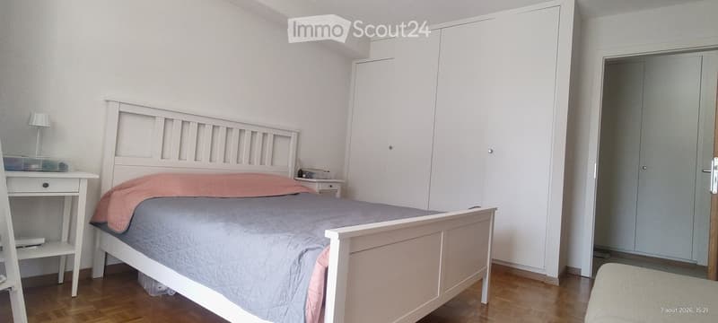 A modern bedroom with a white bed, grey bedding, a nightstand with a lamp, a wardrobe, and a small desk with a chair.