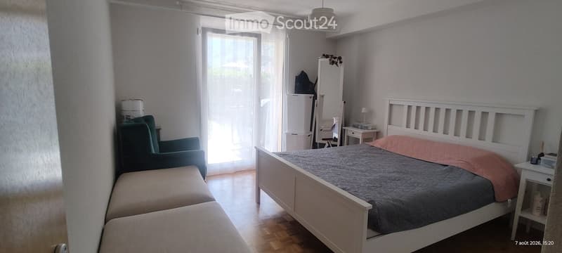 Bright bedroom with wooden flooring, a double bed with grey bedding, a green armchair, a small nightstand, and a mirror on a dresser.