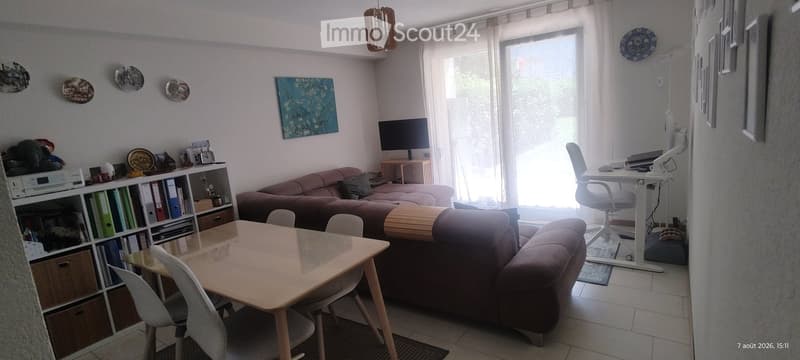 A well-lit living room with a sofa, a coffee table, a dining table with chairs, a bookshelf, a TV screen, and a desk with a chair.