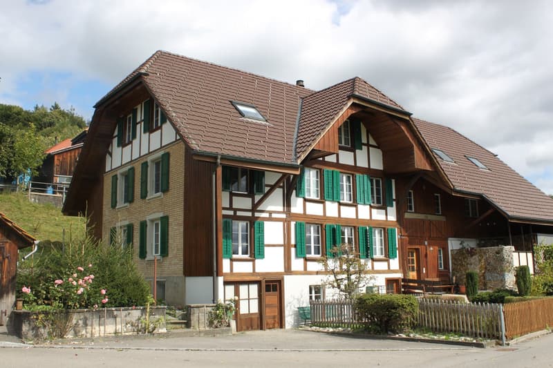 Traditional two-story house with a red-tiled roof, white and brown wooden facade, green shutters, and a small garden area with a wooden fence.