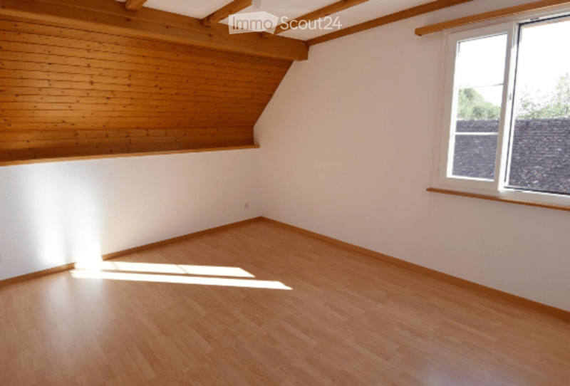 Empty room with wooden flooring and white walls, featuring a slanted wooden ceiling and a window letting in natural light.