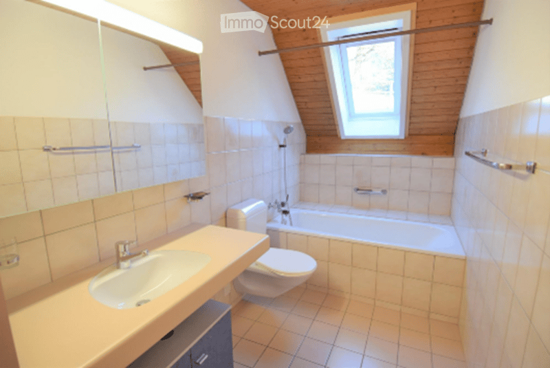 Bright bathroom with wooden ceiling beams, white tiled walls, a sink, toilet, and bathtub. A window provides natural light.