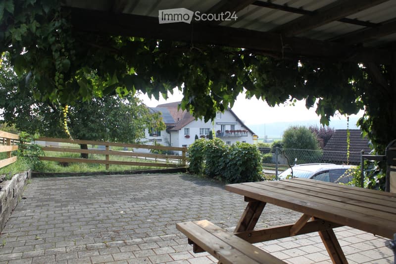 A covered outdoor area with a wooden picnic table, brick pavement, and surrounding greenery, with a view of a house and countryside in the background.