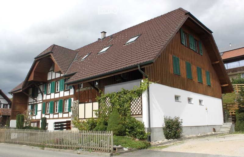 Two-story house with a red-tiled roof, wooden facade, green shutters, and a white lower section. Features a balcony, small garden, and wooden fence.