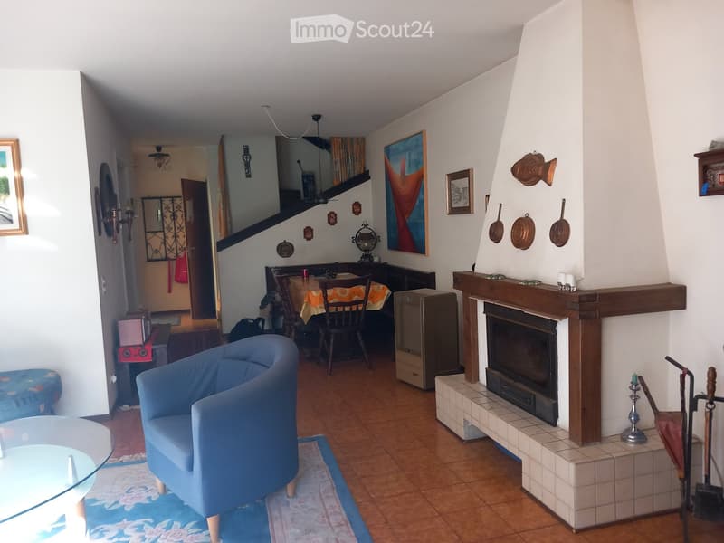 Living room with a fireplace, blue sofa, glass coffee table, framed artwork, and a dining area with a table and chairs.