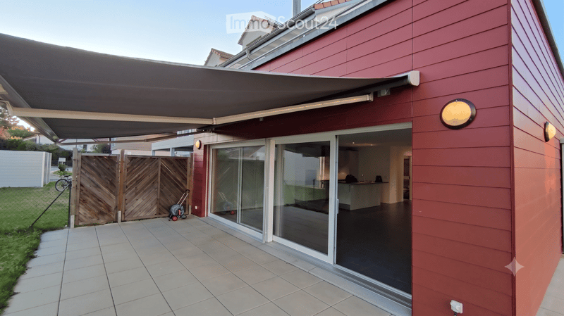 Modern outdoor patio area with a retractable awning, large glass sliding doors leading into a room, tiled floor, wooden gate, and a small grassy area.