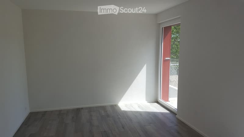 Empty room with wooden flooring and white walls, featuring a large window with a red door leading outside.