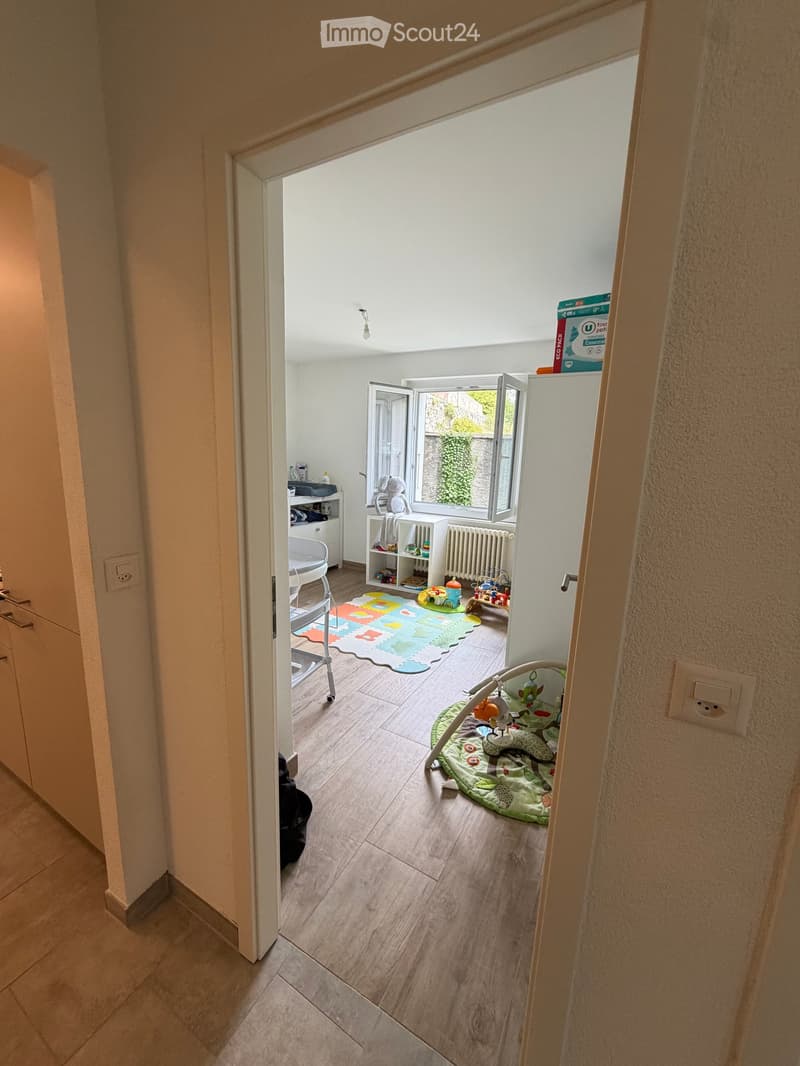 A well-lit children's room with wooden flooring, a colorful play mat, a baby seat, and various toys.