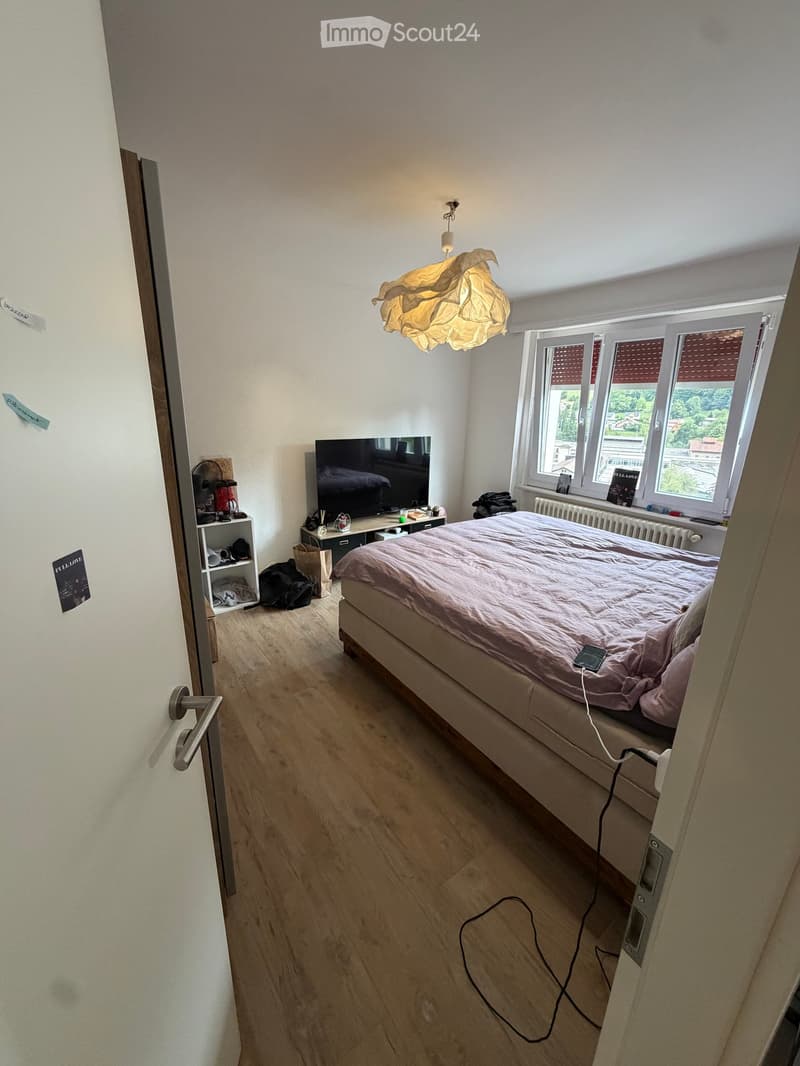 A well-lit bedroom with a large bed, a TV on a stand, a shelf with various items, and a fan near the window. The room has wooden flooring and large windows with blinds.