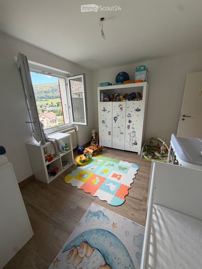 Brightly lit children's room with wooden flooring, large window, colorful play mats, a wardrobe with cartoon designs, shelves with toys, and a baby chair.