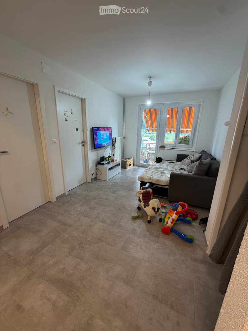 Living room with a sofa, bed, television, small table, and toys on the floor. The room has large windows with blinds and a tiled floor.