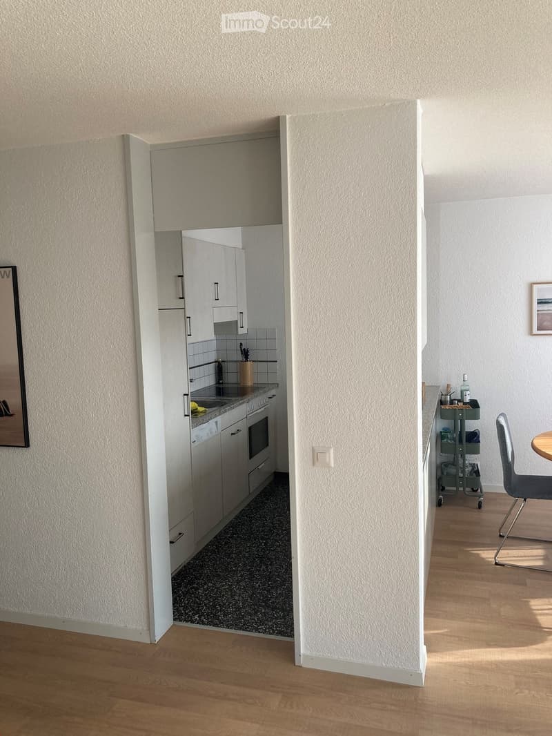 A modern, compact living space with a small kitchen area visible through an open doorway. The kitchen features white cabinets, a sink, and an oven. The living area includes a wooden floor, a chair, a small table, and a cart with various items.