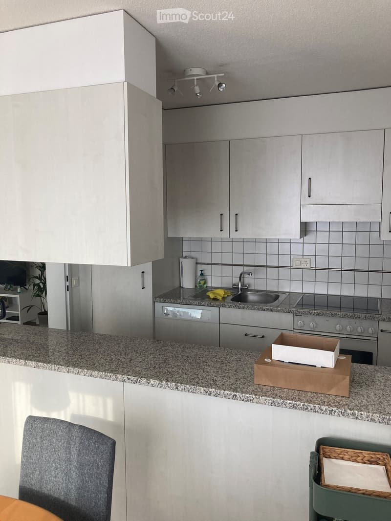 Modern kitchen with white cabinets, granite countertop, sink, oven, dishwasher, and a small chair near the counter.