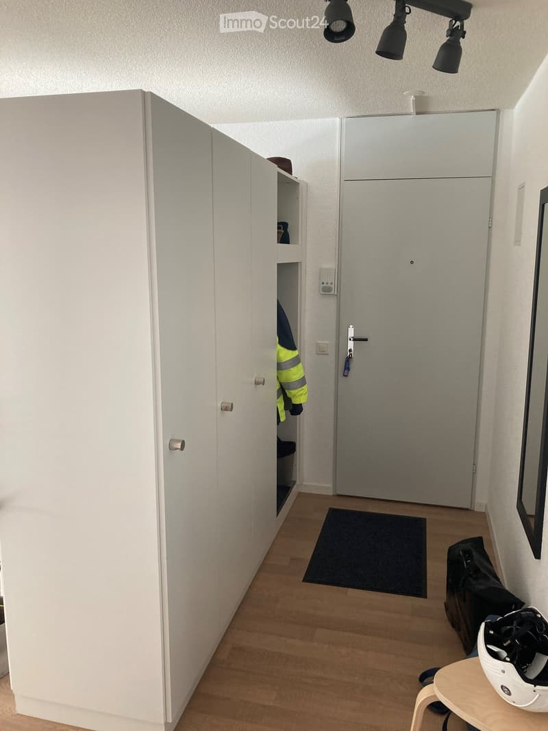A small, modern hallway with a wooden floor, white walls, and a built-in wardrobe. A helmet and backpack are placed on a small wooden stool near the door.