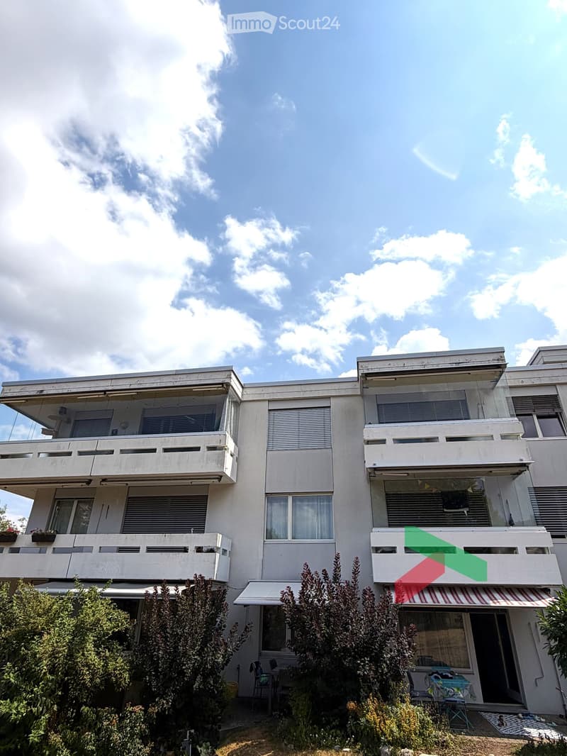Modern multi-story building with balconies, large windows, and a striped awning over the entrance. The building has a clean, minimalist design with white walls and concrete elements.
