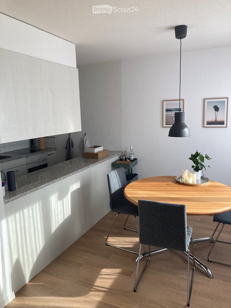Modern kitchen with white cabinets, granite countertop, round wooden dining table with four chairs, pendant light, and decorative items including a plant and candles.