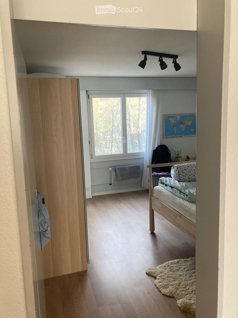 Bright bedroom with wooden flooring, large window with white curtains, bed with white bedding, fluffy white rug, radiator, world map on wall, small plant, and backpack.