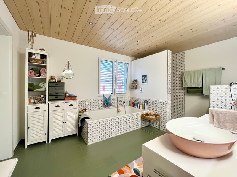 Bathroom with green tiled floor, white cabinets, a bathtub with patterned tiles, a sink, and a shelf with towels and toiletries.