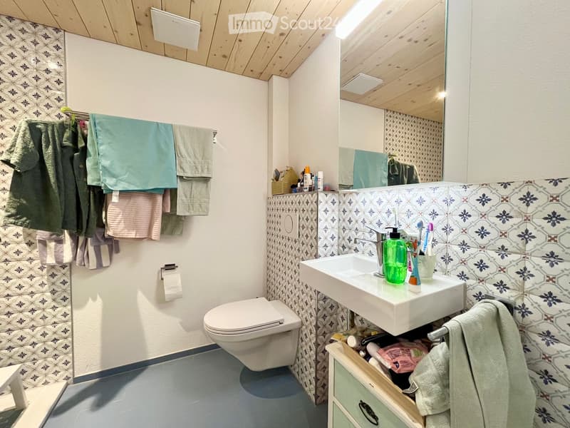 Modern bathroom with a toilet, sink, mirror, and patterned tiles. Towels are neatly hung on a rack, and toiletries are placed on the sink counter.