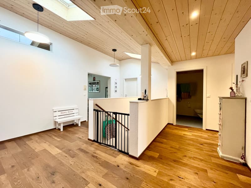 Bright hallway with wooden flooring, white walls, and a staircase with a safety gate. Features pendant lights, a small bench, and a view into a bathroom and another room.