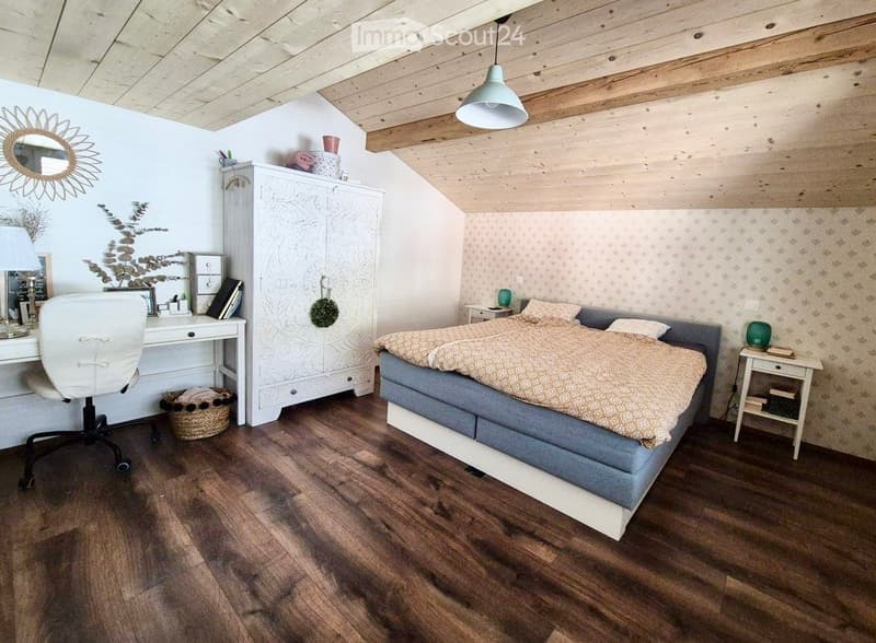 Cozy bedroom with wooden flooring, slanted wooden ceiling, and modern decor. Features a large bed with a grey headboard and beige bedding, a white desk with a chair, a white cabinet, and decorative items including a mirror and a wicker basket.