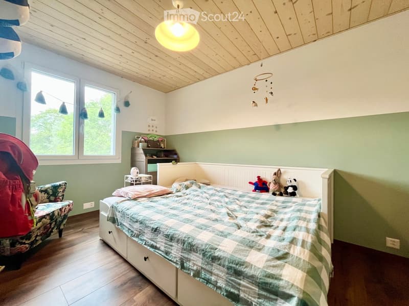 Cozy children's bedroom with a large bed, colorful quilt, stuffed toys, a small table, and a hanging mobile. The room features wooden flooring, a slanted ceiling, and large windows with a view of greenery.