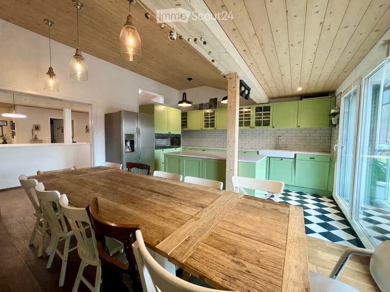 Open-plan dining and kitchen area with a large wooden dining table surrounded by white and wooden chairs, green cabinets, a stainless steel refrigerator, and a white tiled backsplash.