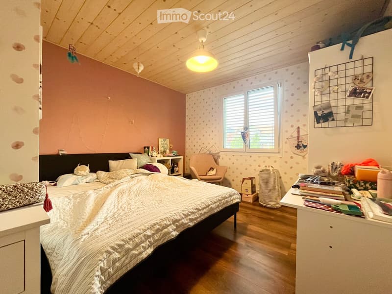 Cozy bedroom with wooden floors, a large bed with pillows and blankets, a nightstand, a chair, a table with various items, and a hanging lamp.