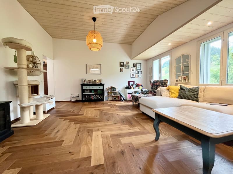 Bright living room with wooden flooring, white walls, and large windows. Features a white sectional sofa with colorful cushions, a wooden coffee table, a tall cat tree, a bookshelf, and a patterned armchair.