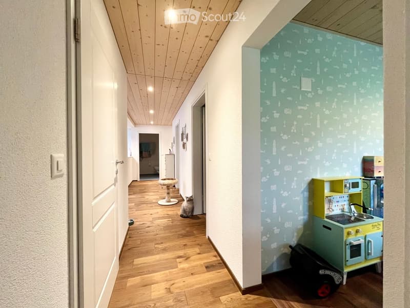 A narrow hallway with wooden flooring, white walls, and a cat sitting near a cat tree. A small play area with a toy kitchen set is visible on the right side.