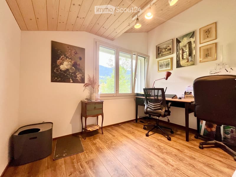 Cozy study room with wooden flooring, large windows, a desk with a chair, a small table with a vase, and several picture frames on the walls.