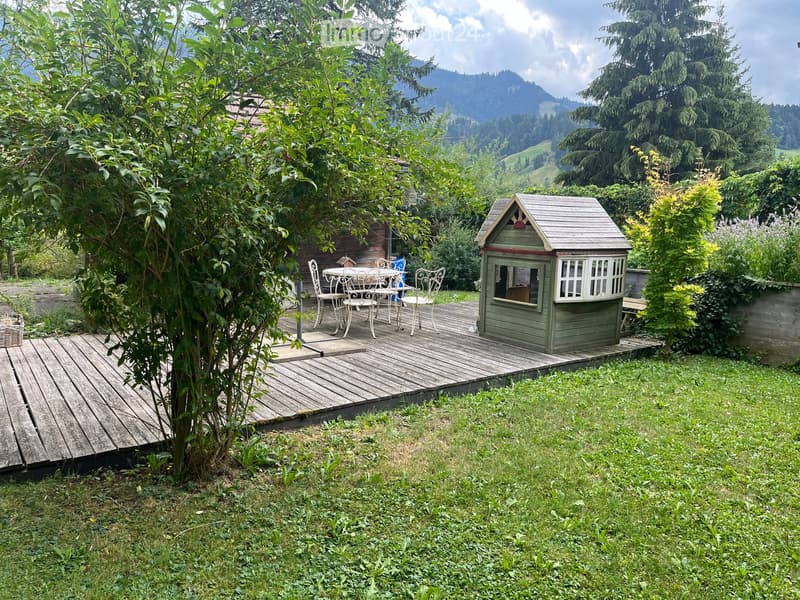A wooden deck with a small table and chairs, a playhouse, and surrounding greenery with a scenic mountain view in the background.