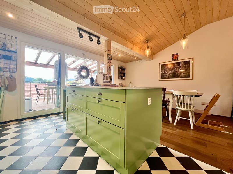Bright kitchen with a green island, checkered floor, wooden ceiling, and large windows opening to a balcony. Includes a dining area with chairs and a table.
