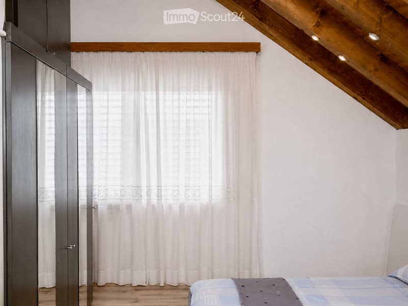 Cozy attic bedroom with wooden slanted ceilings, white walls, and a large window with white curtains. Features a bed with a blue and grey checkered duvet and a modern grey wardrobe.