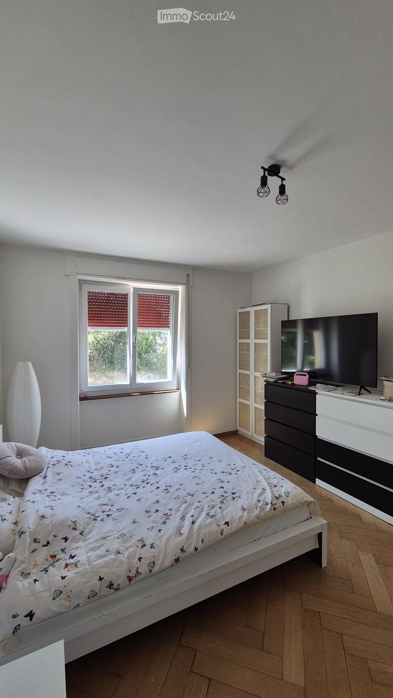 Bright bedroom with wooden flooring, a white bed with butterfly-patterned bedding, a nightstand with a lamp, a wardrobe, and a television on a black and white stand.