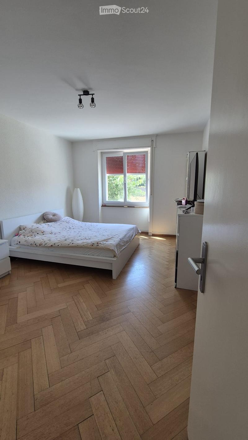 Bright bedroom with wooden herringbone flooring, white walls, a large window with blinds, a single bed with bedding, a nightstand, and a decorative vase near the window.