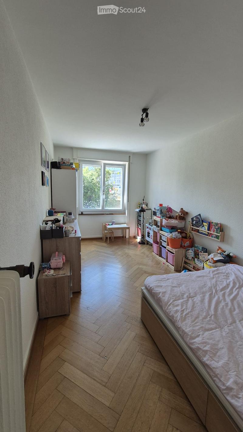 Bright bedroom with wooden herringbone flooring, large window, bed with light pink bedding, nightstand, small table with chairs, storage unit, bookshelf, and various toys and books.
