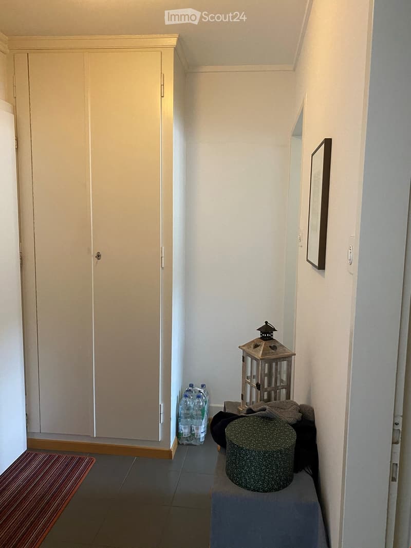 A narrow hallway with a wooden door on the left, a small bench with a stool, a lantern, and a picture frame on the right.