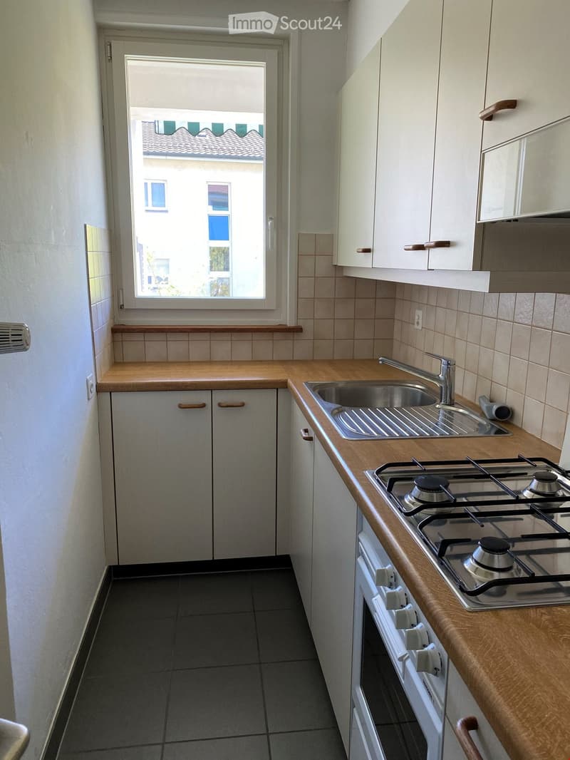 Modern kitchen with light wooden countertops, white cabinets, a gas stove, and a sink. The space features tiled walls and a window offering a view of the building outside.