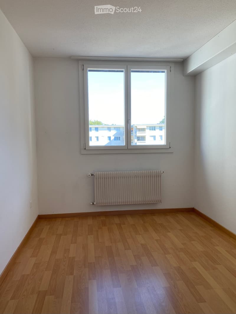 Empty room with wooden flooring, white walls, a window with white frame, and a radiator below the window.