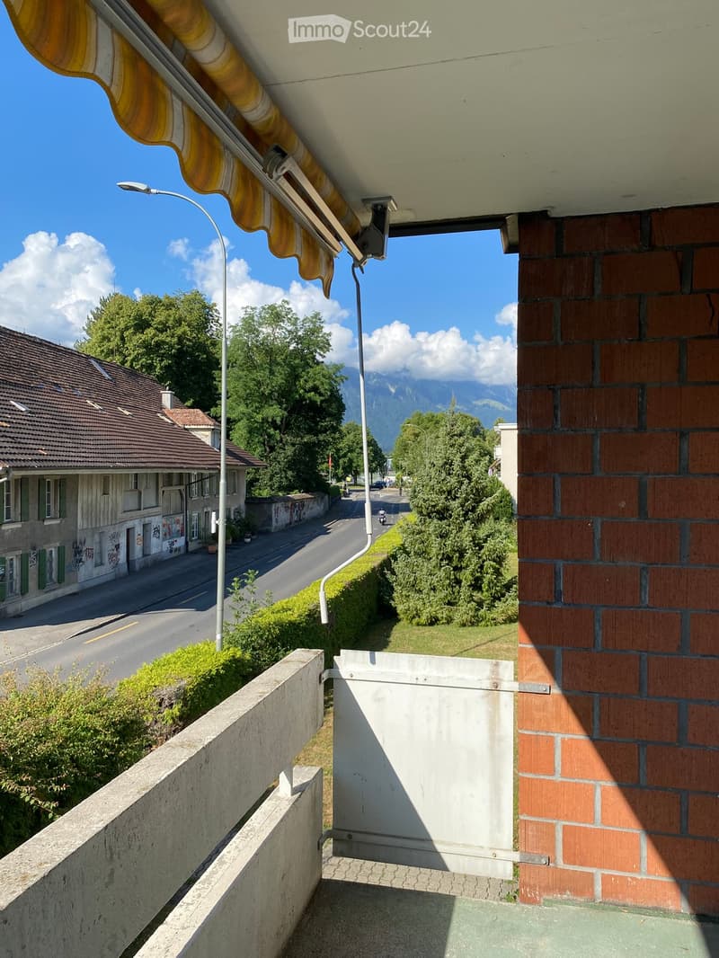 Balcony with a view of a street, houses, and mountains in the background. Features a concrete railing, a brick wall, and a yellow and white awning.
