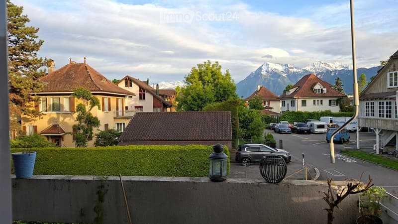 View from a balcony showing a street with parked cars, houses with brown roofs, and snow-capped mountains in the background.