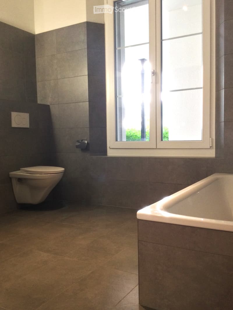 Modern bathroom with a bathtub, toilet, tiled walls, and a large window letting in natural light.